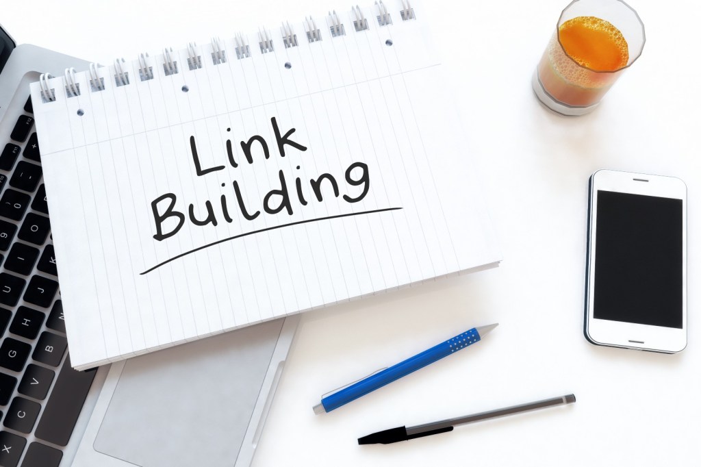 Notebook with 'Link Building' written on it placed on a laptop keyboard, alongside a pen, a smartphone, and a glass of orange juice on a white desk.